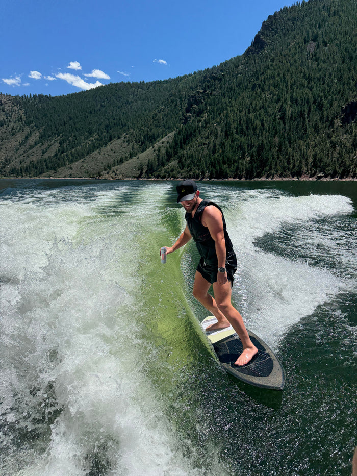 A smiling man in a black hat and life vest wakesurfs on a vibrant green lake under a clear blue sky, with forested mountains in the background.