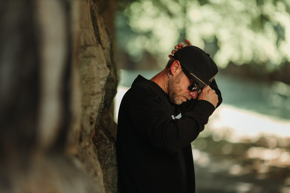 A man in a black hoodie and black Suay cap leans against a stone wall, looking down, with a soft, blurred natural background.