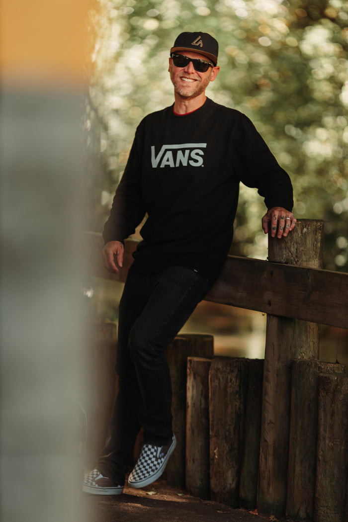 A smiling man wearing a black Suay Apex hat with a gold LA logo and a black Vans sweatshirt leans on a wooden fence in a warm outdoor setting.
