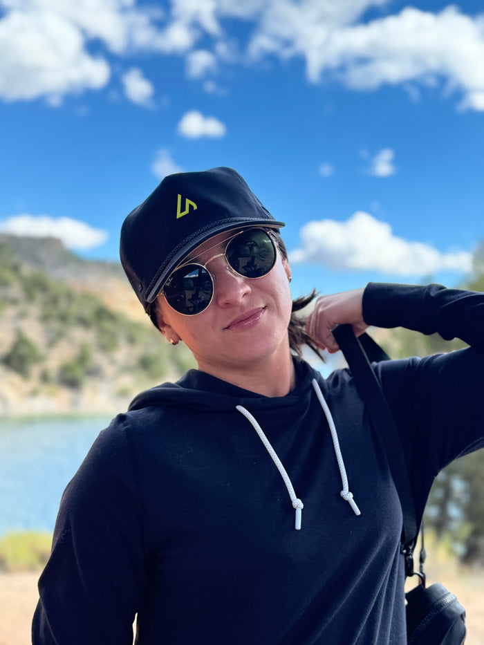 A young woman wearing a black Suay cap with a neon logo and sunglasses, smiling slightly outdoors against a clear blue sky, white clouds, and a body of water.