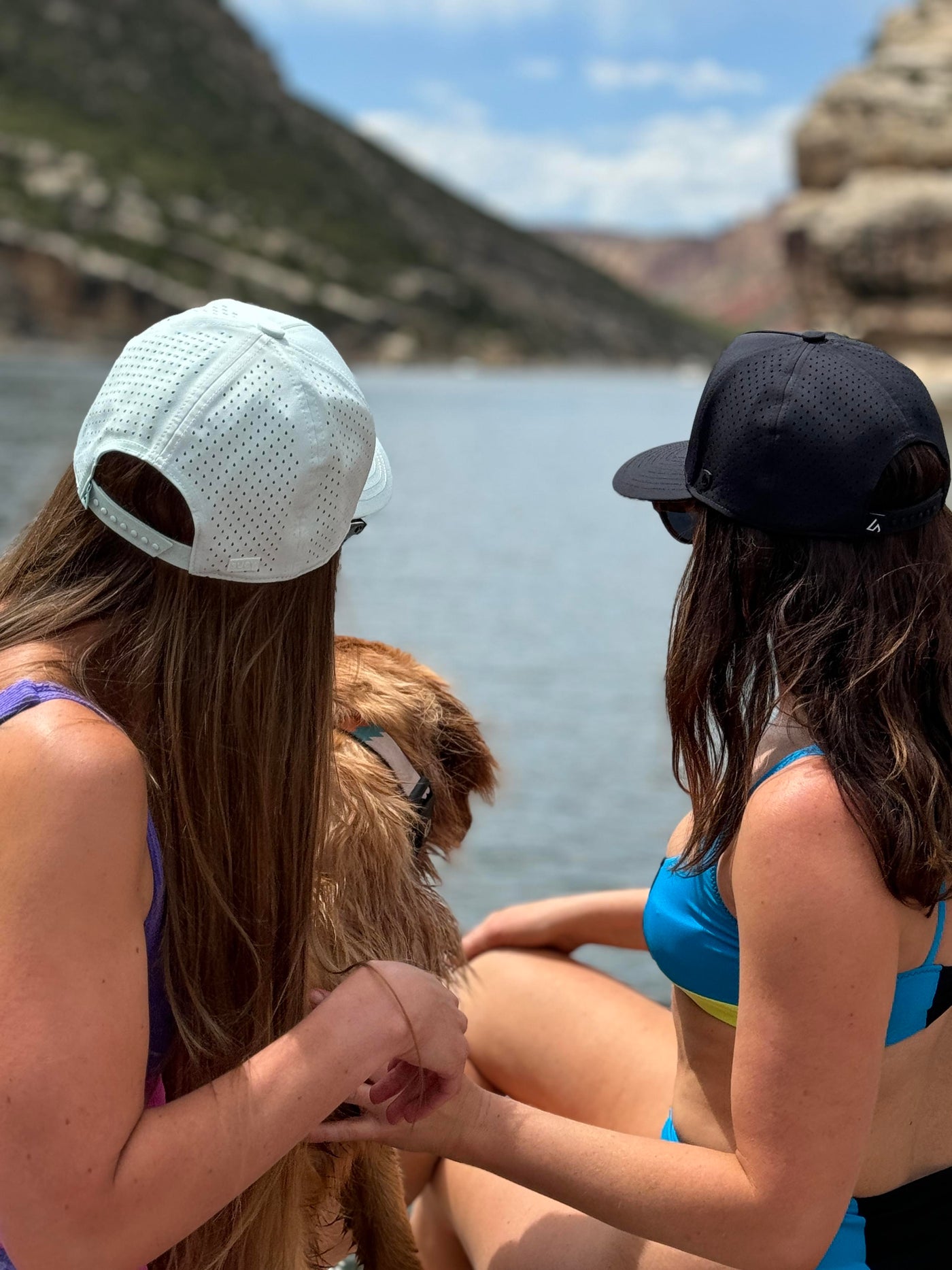 Two women wearing Suay Apex hats, one mint and one black, and a dog, sit by a serene lake with mountains in the background, looking out at the water.