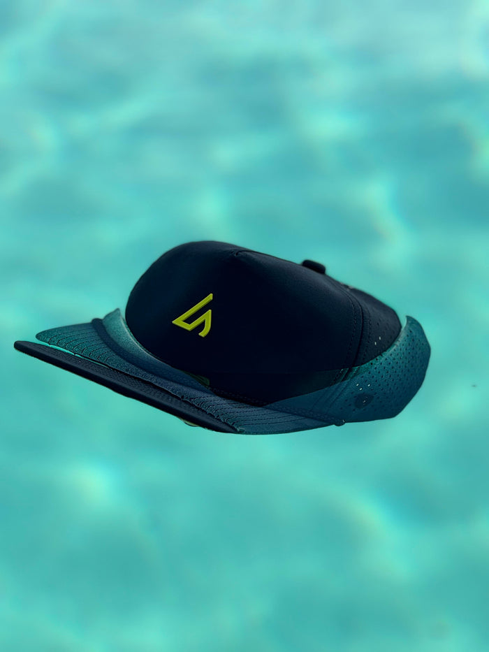 A black Suay Apex hat with a vibrant yellow logo floats effortlessly on the calm, clear turquoise water, showcasing its water-friendly design.