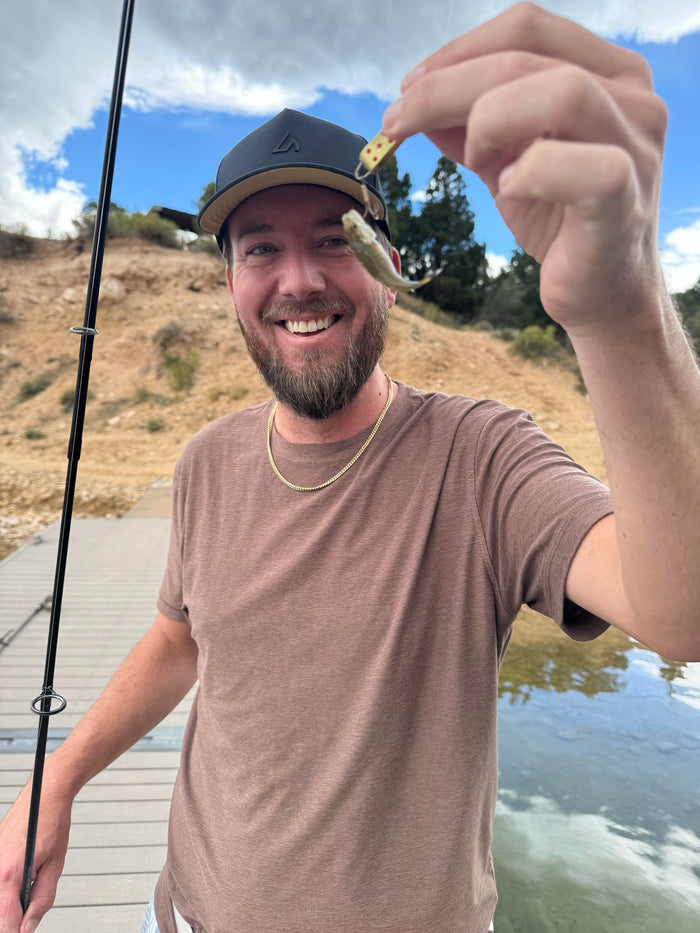 A smiling man wearing a dark Suay cap holds up a tiny fish and a fishing lure while standing on a dock by a lake with hills in the background.