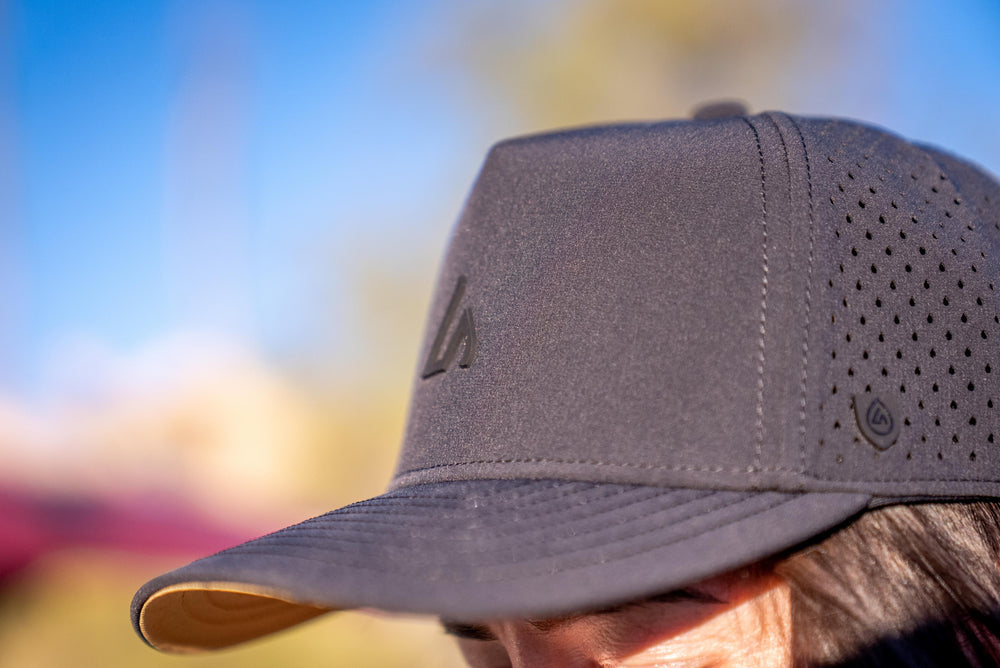 Close-up of a person wearing a dark grey Suay Apex hat outdoors, featuring the front logo, perforated mesh panels, and a blurred sky background.