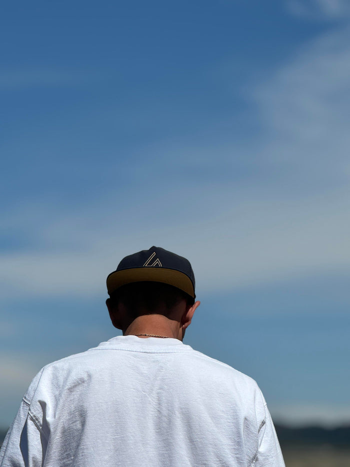 A person wearing a black Suay Apex hat with a gold brim and logo is seen from behind, looking out at a bright blue sky.