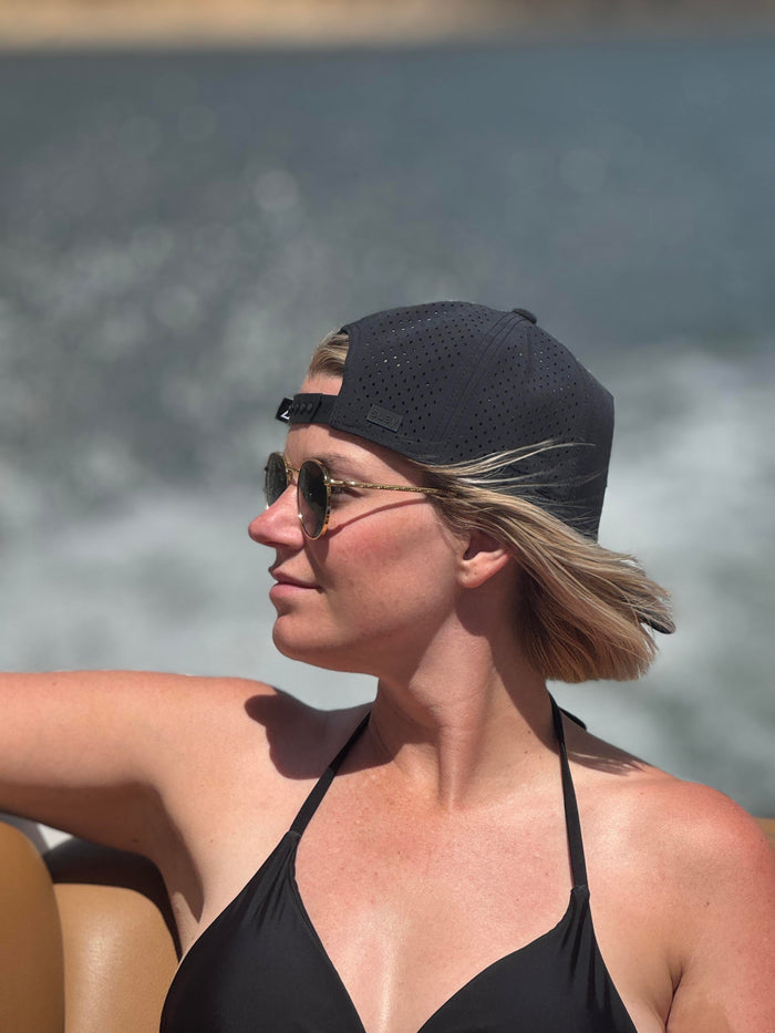 A woman wearing a black Suay Apex hat and sunglasses looks to the left while on a boat, with blurred water and sky in the background.
