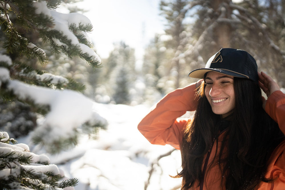 A smiling woman with dark hair and freckles wears a black Suay hat and an orange jacket in a sunlit snowy forest, adjusting her hat.