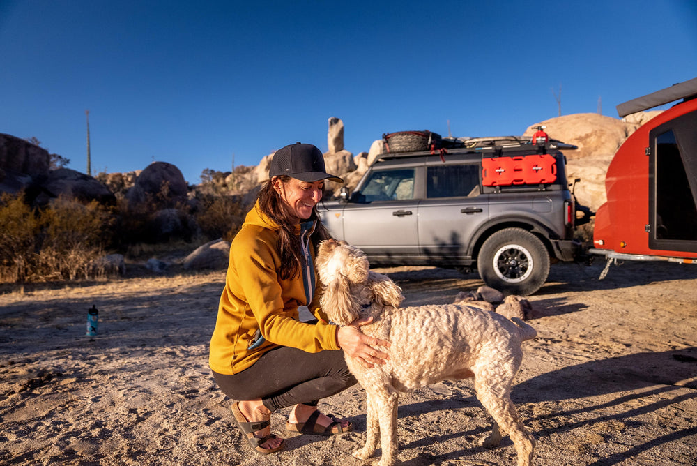 A smiling woman in a yellow hoodie and black Suay Apex hat crouches, affectionately petting a fluffy light-colored dog in a sunny rocky desert campsite.