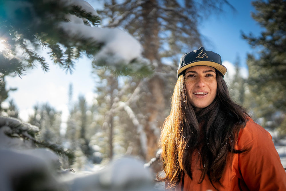 A smiling woman wearing an orange jacket and a black Suay cap stands in a brightly lit, snowy forest with sun flares and blurred pine branches.