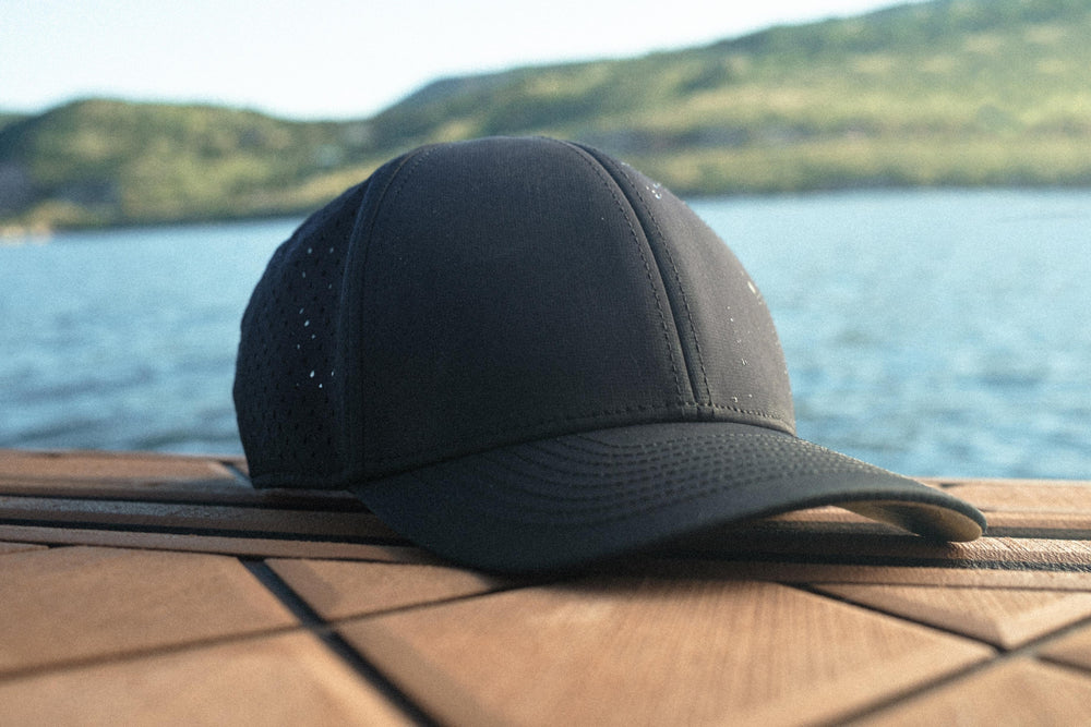 A black Suay Apex hat sits on a textured wooden surface, with a blurred backdrop of a calm lake and distant green mountains.