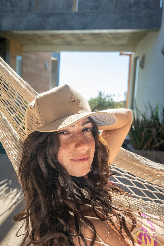 A smiling woman with freckles relaxes in a woven hammock, wearing a light tan Suay Apex cap, looking directly at the viewer on a sunny day.
