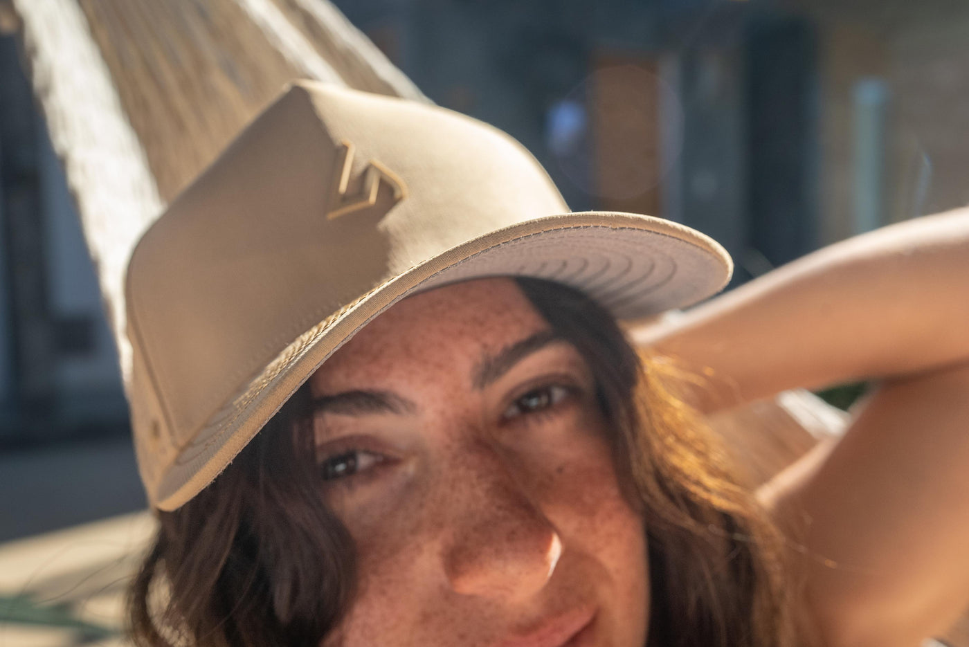 A close-up, low-angle shot of a smiling woman with freckles wearing a camel Suay Apex hat, looking towards the camera in bright natural light.