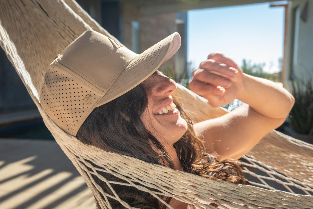 A smiling woman wearing a dune Suay Apex hat relaxes in a sunlit hammock outdoors, embodying a joyful summer vibe with natural lighting.