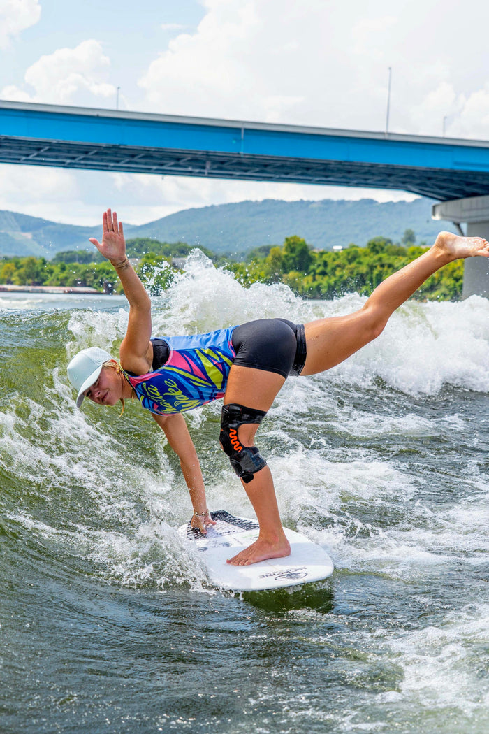 A woman in a colorful top and black shorts expertly balances on a wakesurf board, wearing a light mint green hat, with a blue bridge and green mountains in the background.