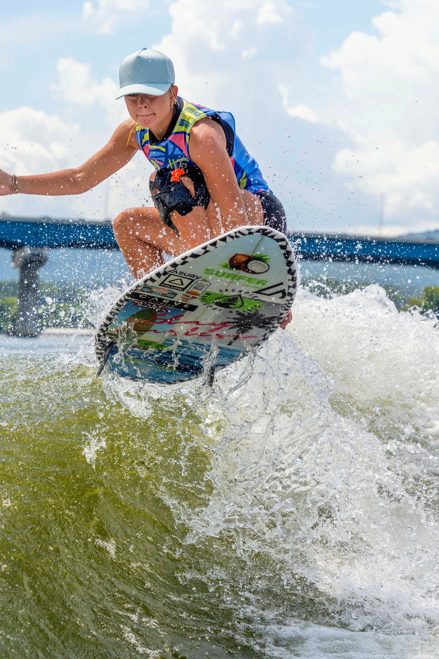 A young woman in an Apex Mint hat wakesurfing on a bright sunny day, captured mid-action with dynamic water spray, set against a blue sky and distant bridge.
