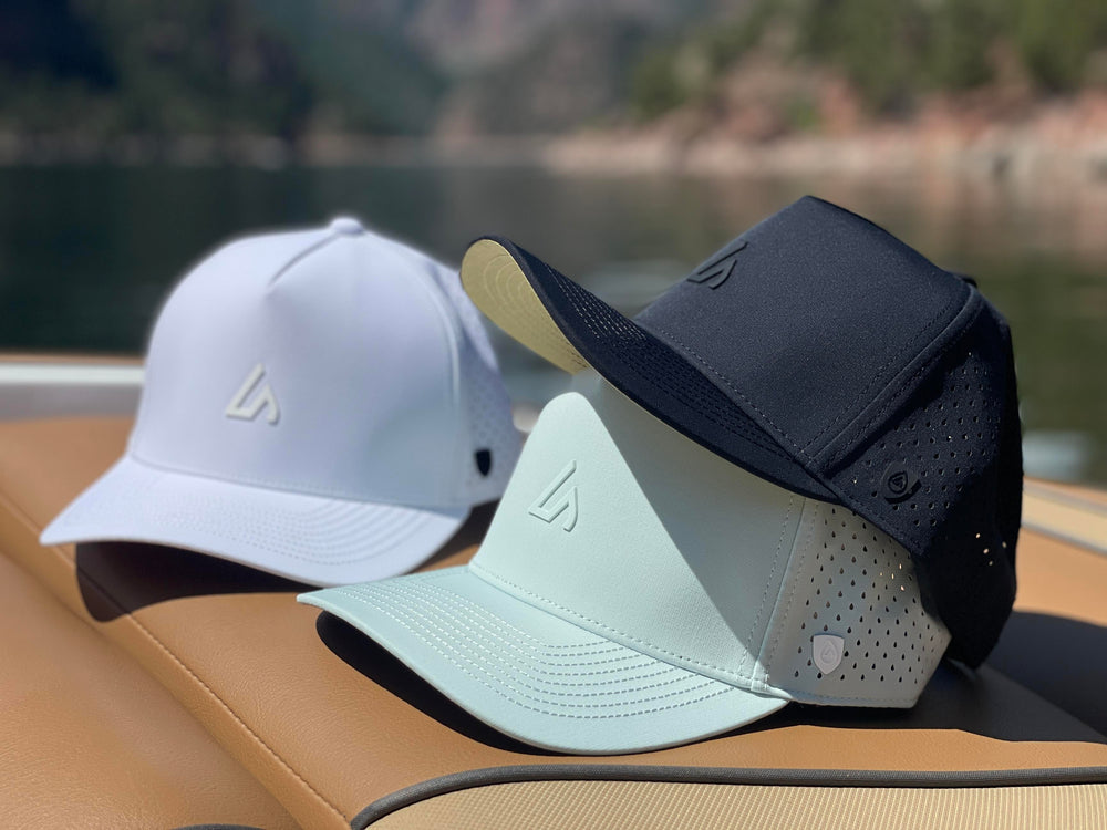 Three premium Suay Apex hats in white, mint, and black, displayed on a boat seat with a blurred lake and mountains in the background under bright sun.
