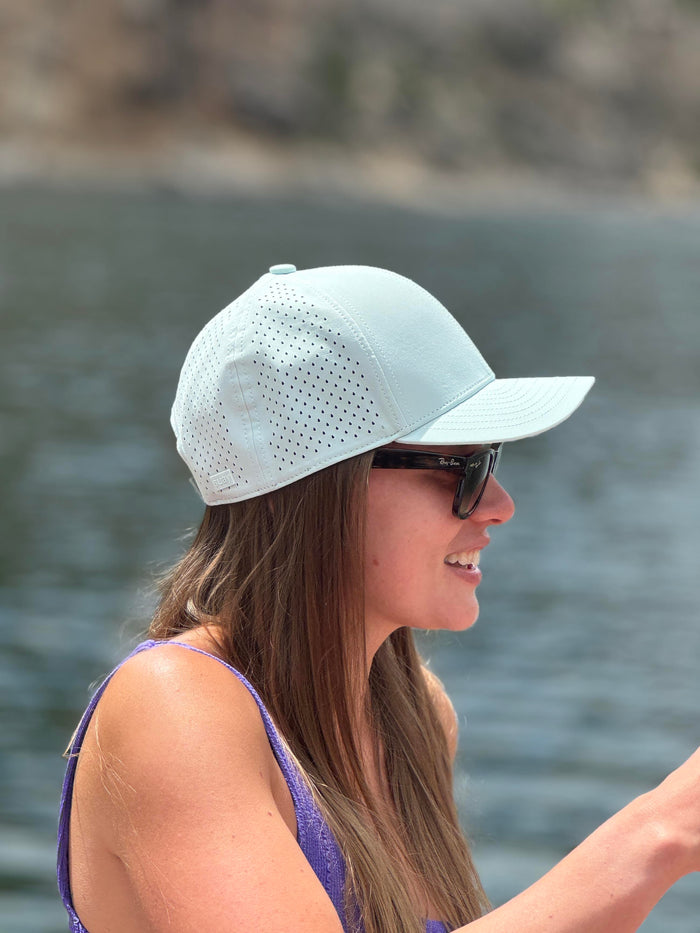 A woman wearing a light mint perforated baseball cap and sunglasses smiles, looking right with a blurred lake and distant shore in the background.