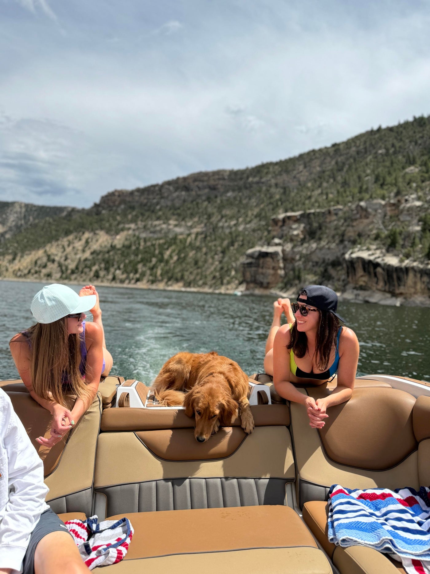 Two women and a golden retriever relax on a boat amidst a scenic lake and mountainous landscape under a bright, cloudy sky.