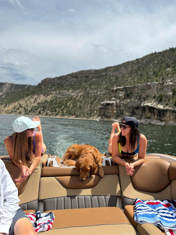 Two women and a golden retriever relax on a boat amidst a scenic lake and mountainous landscape under a bright, cloudy sky.