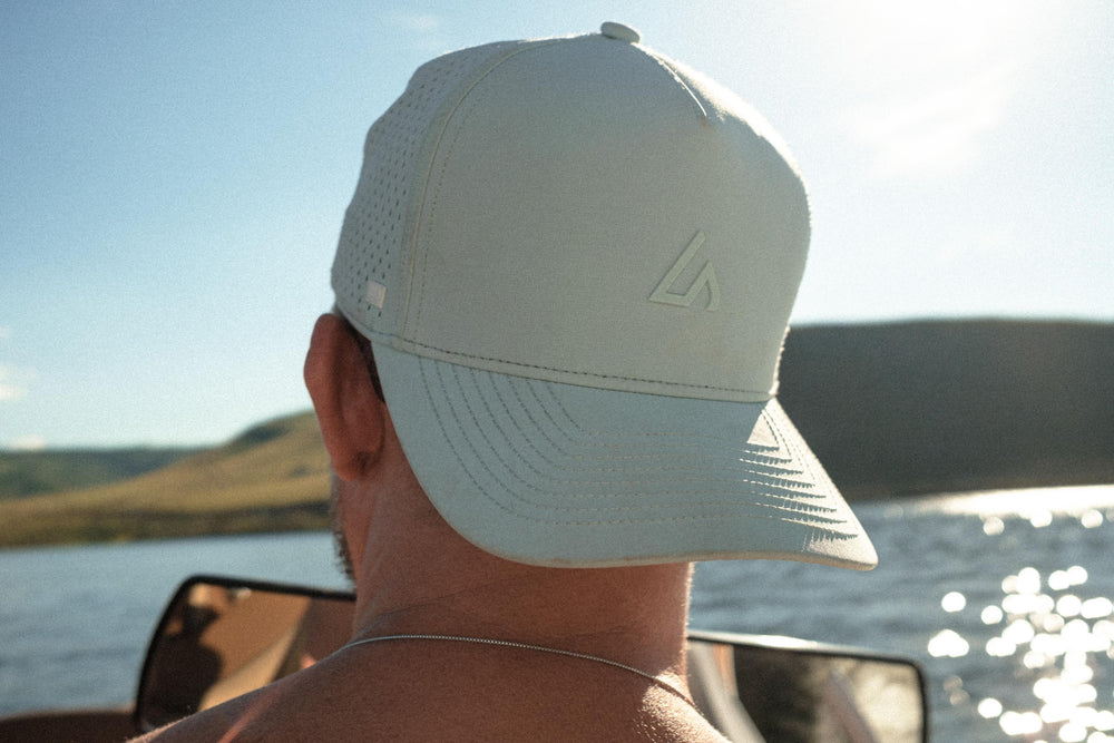 Rear view of a person wearing a light mint performance hat on a sunny day outdoors, looking out over a lake with hills.