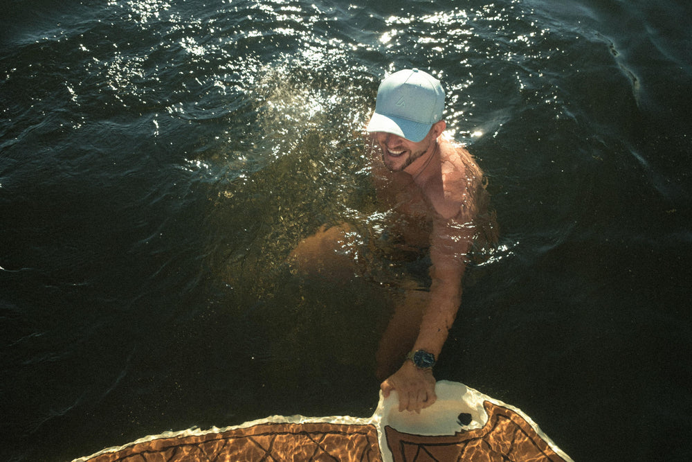A smiling man wearing a light blue Suay cap is submerged in dark water, with sunlight sparkling around him as he holds a boat edge.