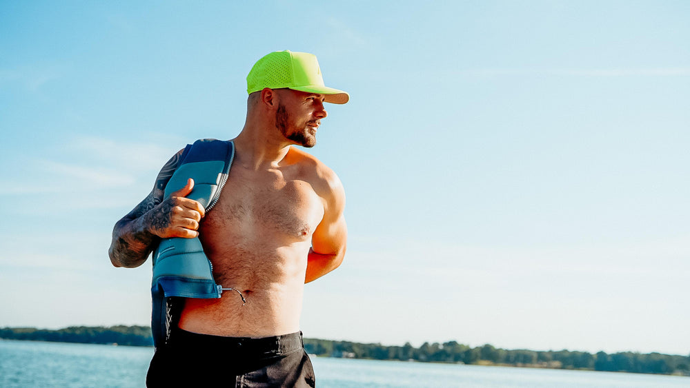 A shirtless man with tattoos and a beard, wearing a bright green mesh cap, holding a blue life vest, stands by a sunny lake.