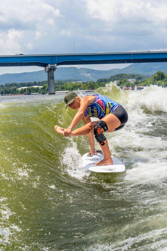A young woman in a green Suay Apex hat wake surfing with impressive balance on a vibrant green river, featuring a blue bridge and forested mountains in the background.