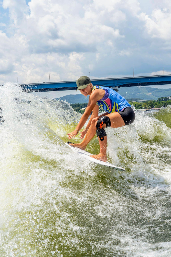 A female wakesurfer wearing a moss green Suay cap and colorful top carves a wave on a sunny day with a blue bridge and mountains in the background.