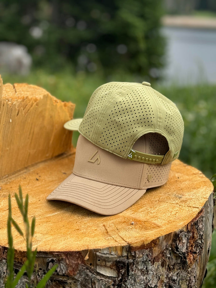 Two performance caps, one moss green and one beige, stacked on a textured tree stump in a lush outdoor setting with blurred greenery and water in the background.