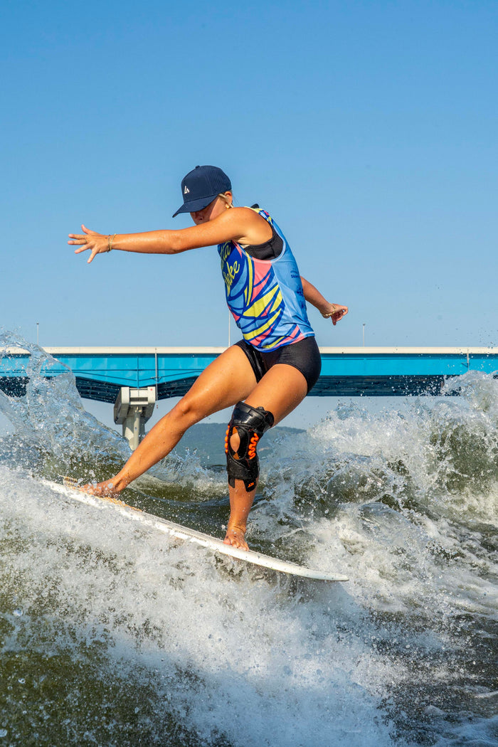 A female wakeboarder in a brightly patterned vest surfs a wake, wearing a navy Suay hat and knee brace under a clear blue sky with a bridge.