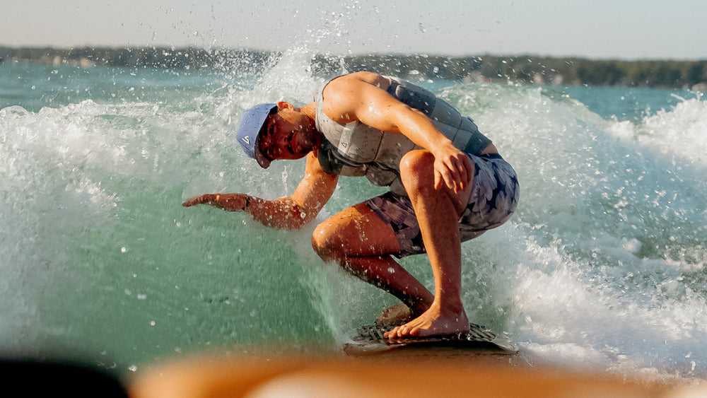 A person wearing a blue baseball cap and life vest rides a wakeboard, carving through a vibrant blue-green wave, with water spraying around them.