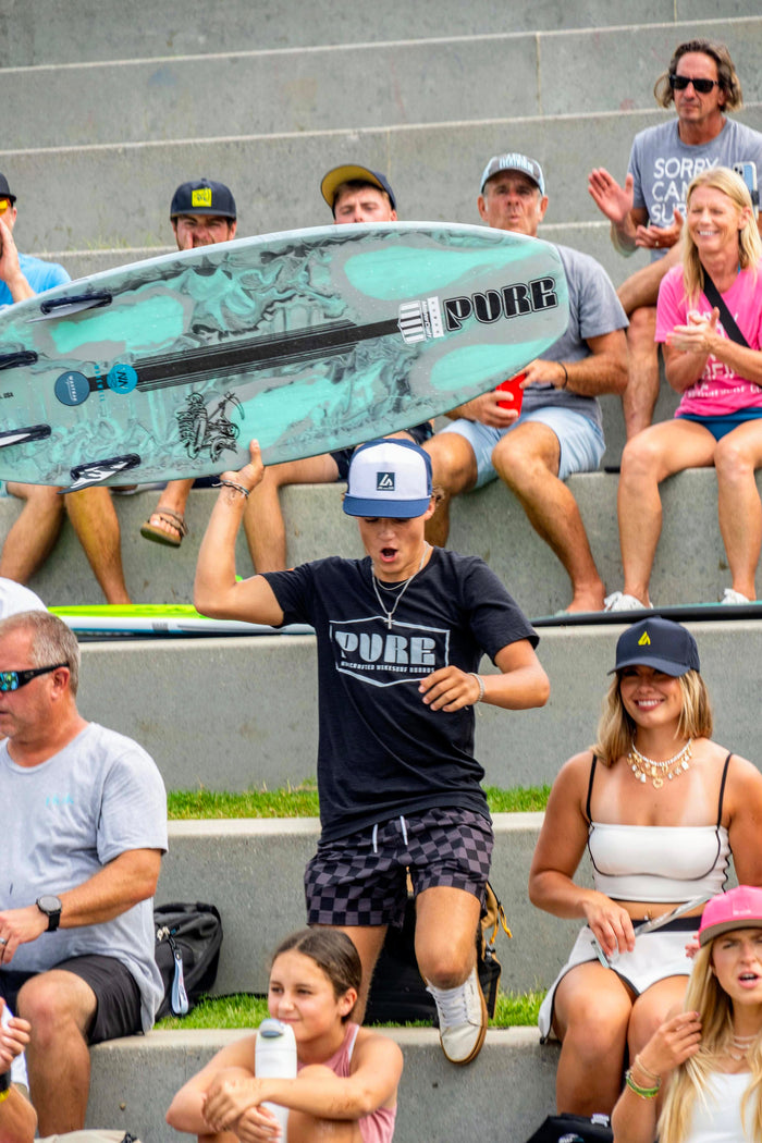 An energetic young man in a Suay hat holds up a wakesurf board while surrounded by a cheering crowd on concrete steps at an outdoor event.