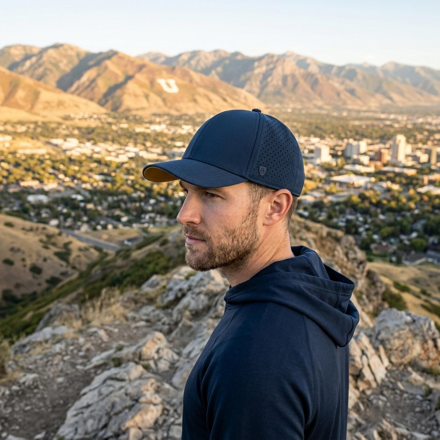 A man in a navy performance cap and hoodie looks left from a rocky mountain overlooking a blurred city and golden mountains at sunset.