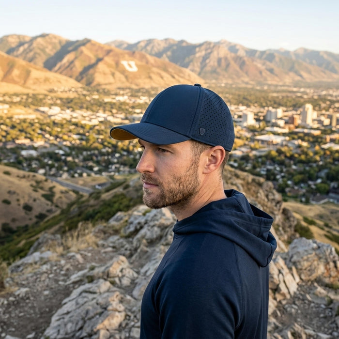 A man in a navy performance cap and hoodie looks left from a rocky mountain overlooking a blurred city and golden mountains at sunset.