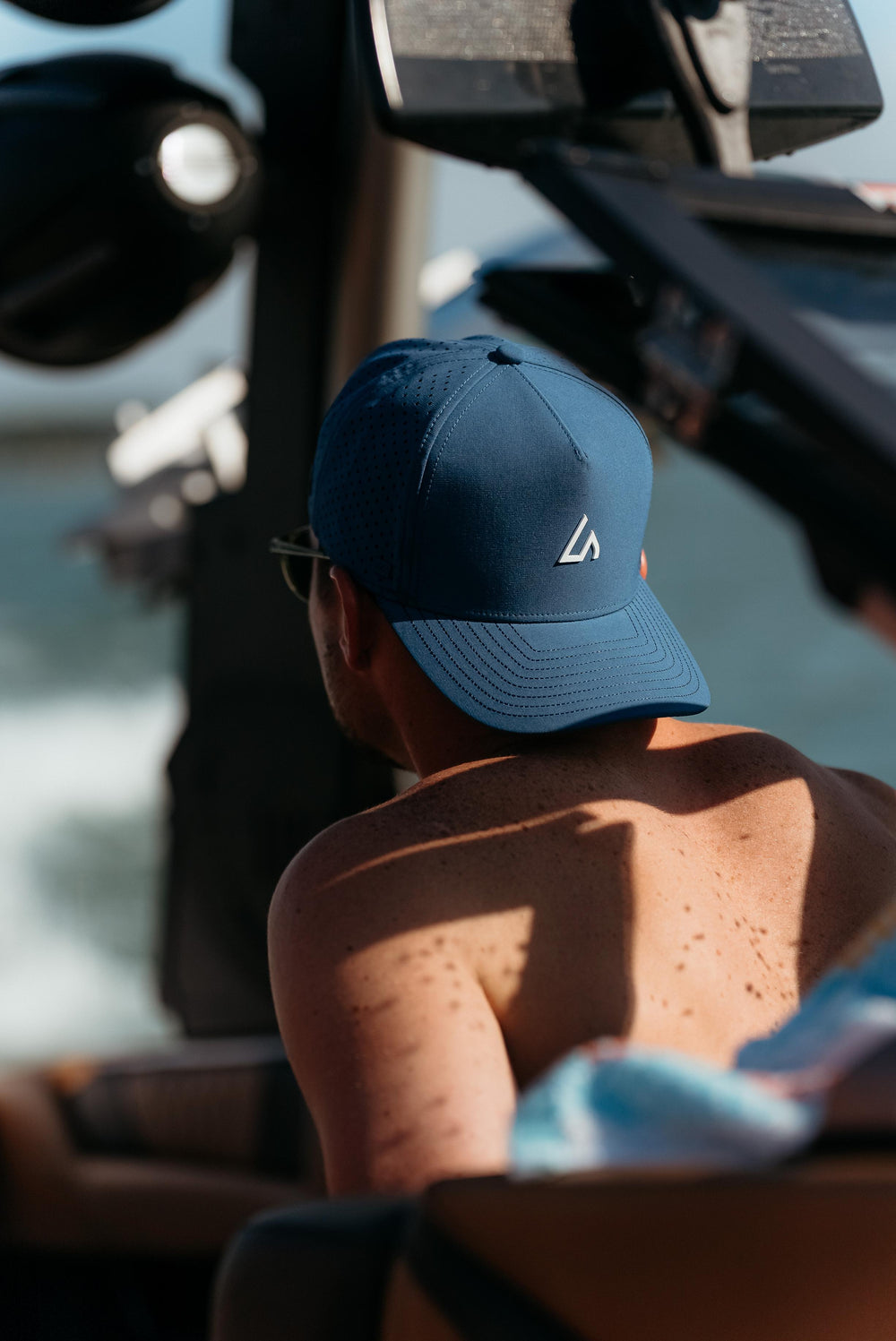 A person from behind wears a navy Suay Apex hat with mesh panels, basking in the sun while enjoying an outdoor, possibly boating, activity.