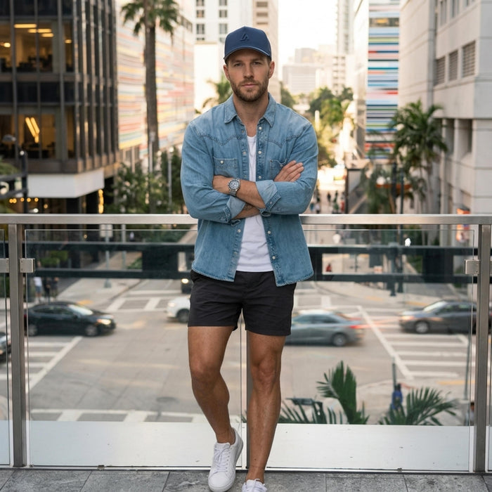 A stylish man in a denim shirt, white tee, and dark shorts wears a navy Suay cap, posing on an urban bridge overlooking a city street.