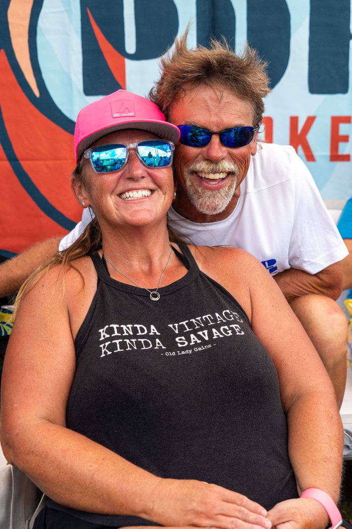 Two smiling adults, a woman in a pink cap and a man with a beard and sunglasses, pose happily at an outdoor event.