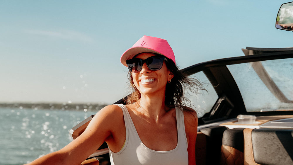 A smiling woman wearing a pink Suay hat and sunglasses enjoys a sunny day on a boat, with bright blue water and sky in the background.