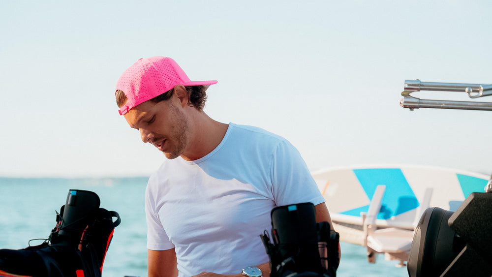 A man wearing a bright pink performance cap and white t-shirt looks down thoughtfully on a sunny day, with blue water and water sports equipment in the background.