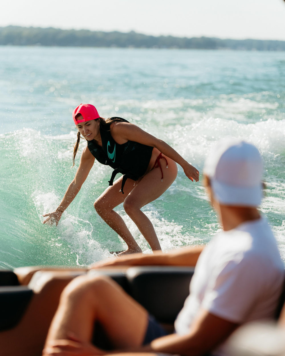 A smiling woman in a neon pink hat and life vest wake surfs on a vibrant blue-green lake, splashing water while an out-of-focus person watches from a boat.