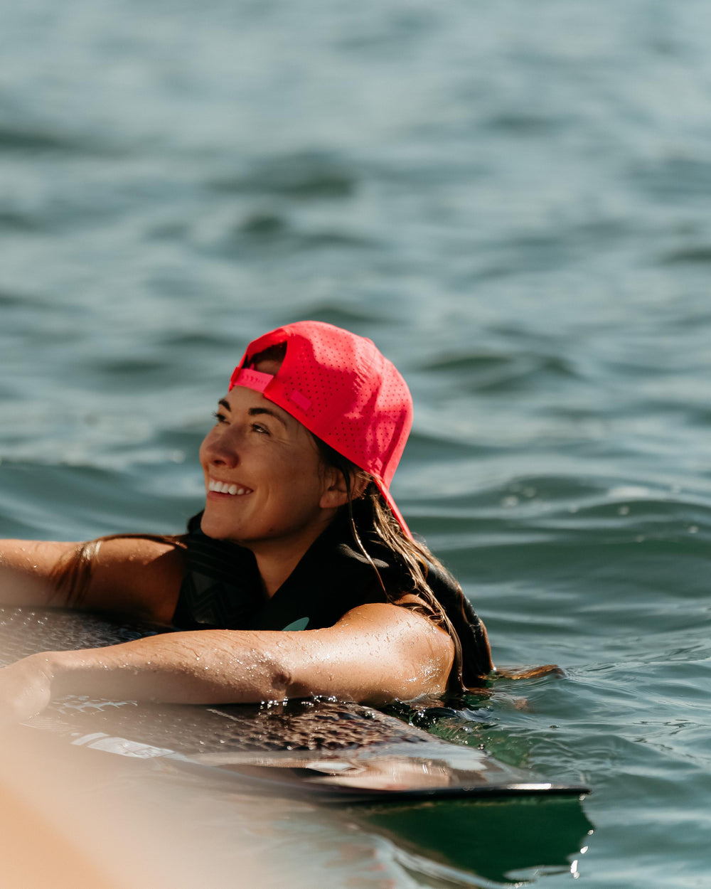 A smiling woman in a vibrant pink Suay hat and life vest, partially submerged in water next to a wakeboard, radiating joy.