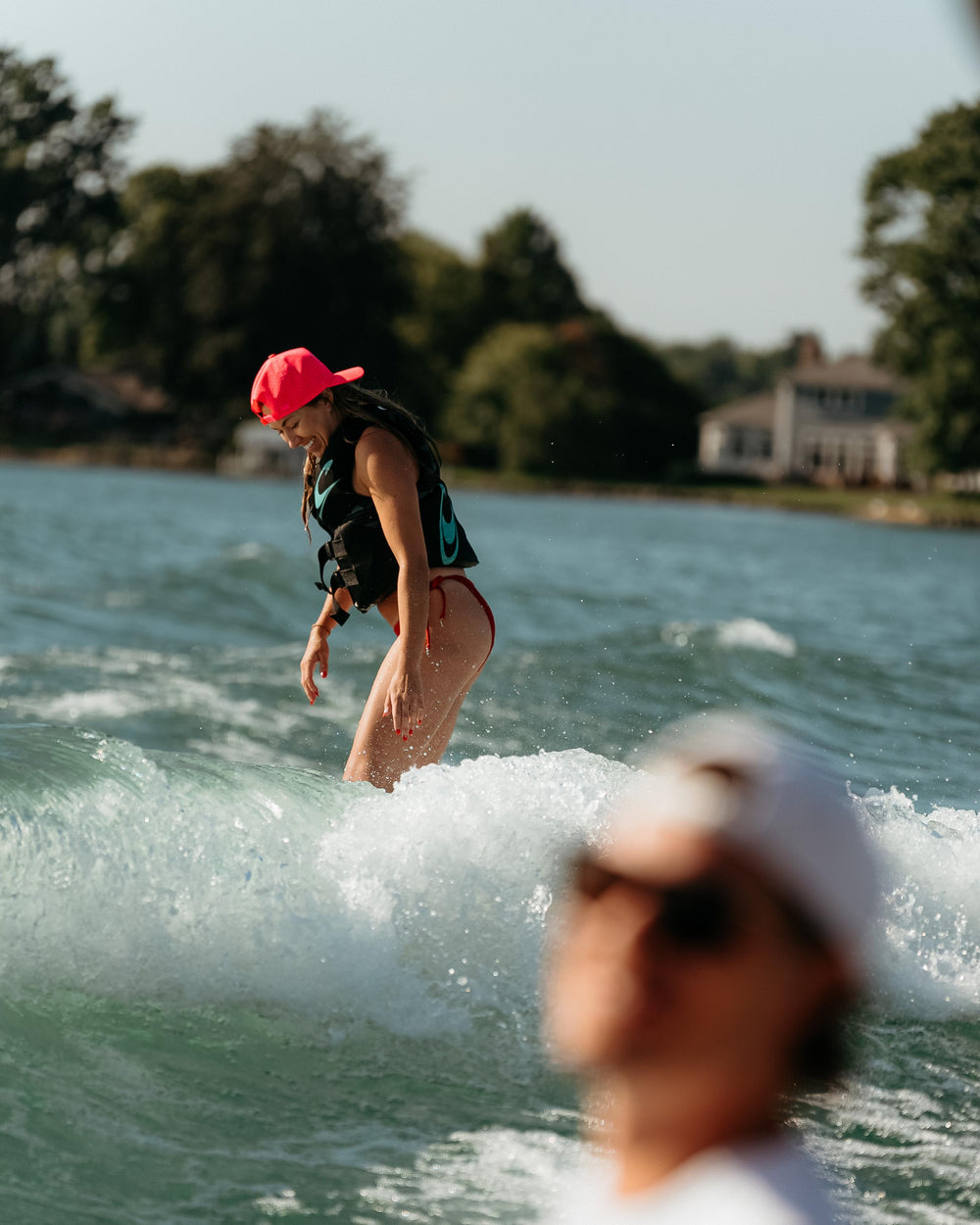 An energetic woman in a bright pink hat and life vest smiles while wake-surfing on clear water with blurred greenery and houses in the background.