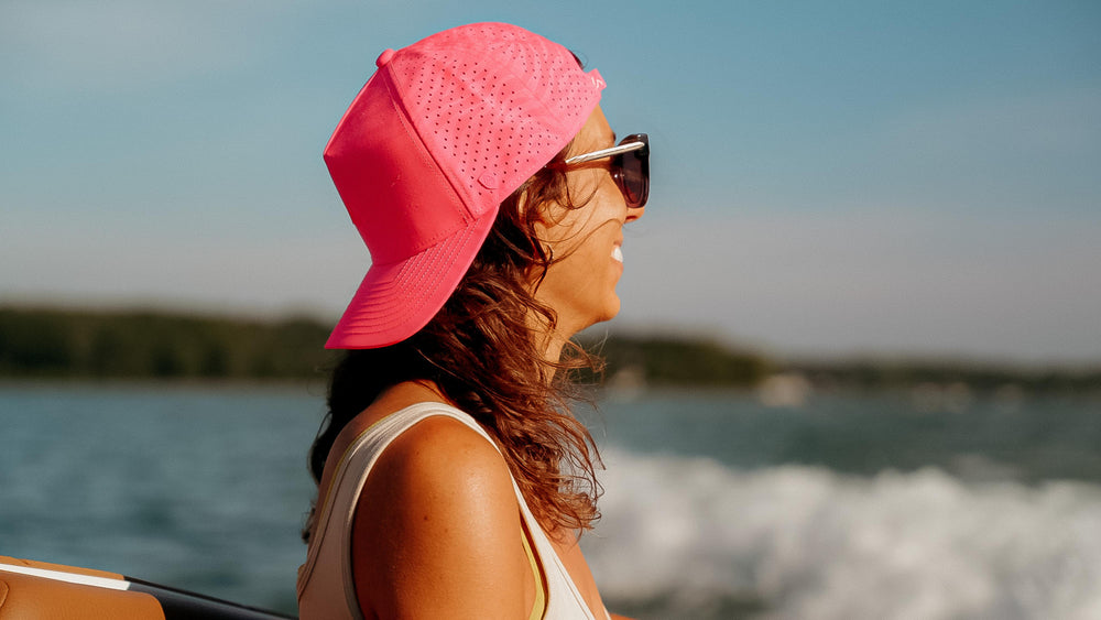 A smiling woman wearing a vibrant pink performance cap and sunglasses on a boat, looking towards the bright blue water with a blurred treeline in the distance.