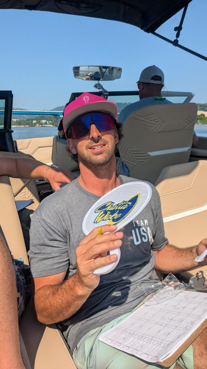 A man in a vibrant pink hat and mirrored sunglasses smiles while holding a small fan on a boat, with a scenic lake and bridge in the background.