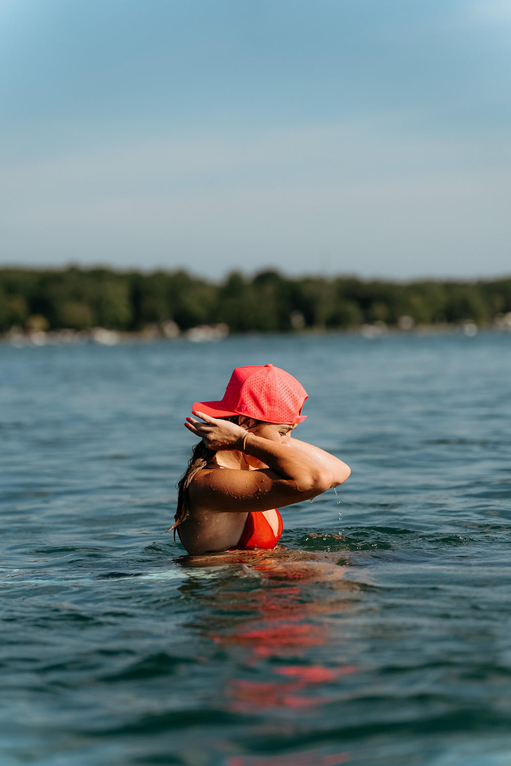 A woman in a red swimsuit and a red Suay cap is partially submerged in a lake, wiping her face with her arm under a bright summer sky.