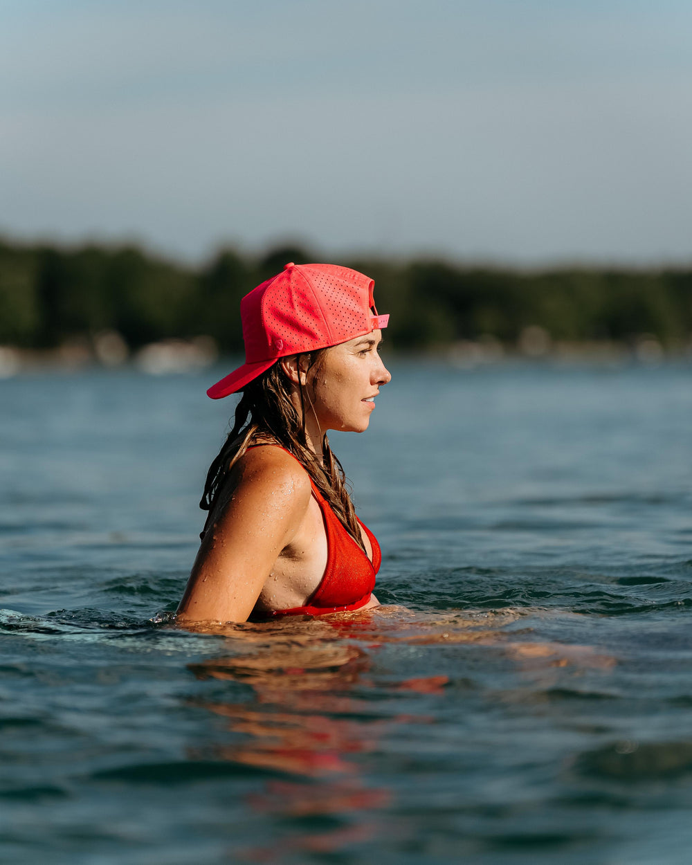 A woman in a red bikini and a bright pink perforated hat wades in blue water, looking right with water droplets on her skin, under golden hour light.
