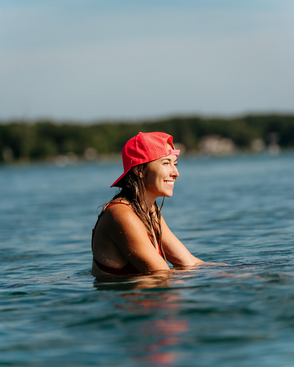 A smiling woman in a bright red Suay Apex hat with wet hair and water droplets on her skin, partially submerged in a serene lake with a blurred natural background.