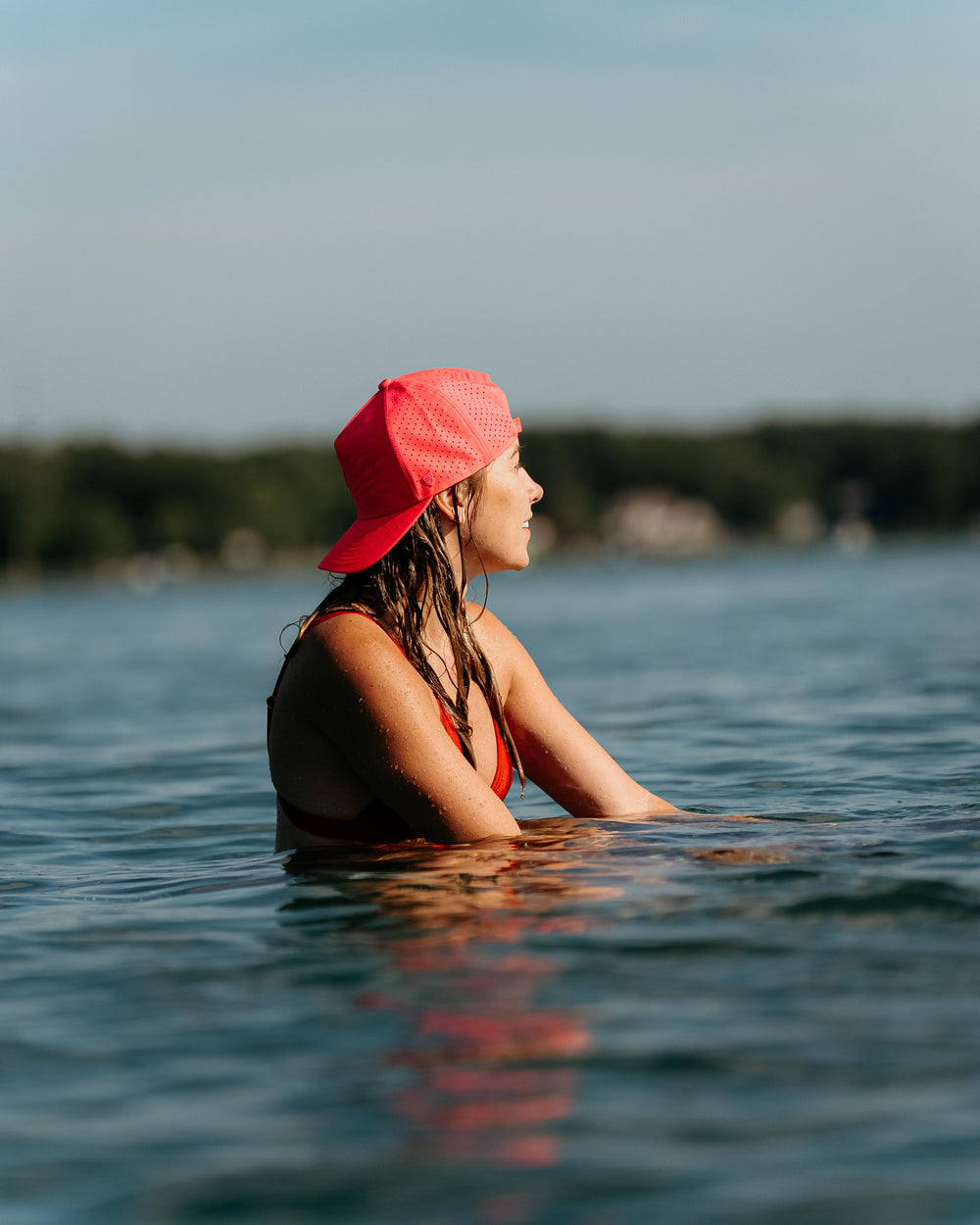 A young woman with wet hair and a bright red perforated Suay Apex cap looks off into the distance while submerged in clear lake water under a sunny sky.