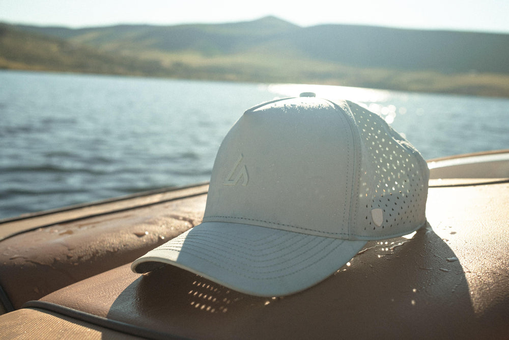 A light-colored performance cap with mesh panels rests on a wet boat deck, overlooking a serene lake and distant mountains under golden sunlight.