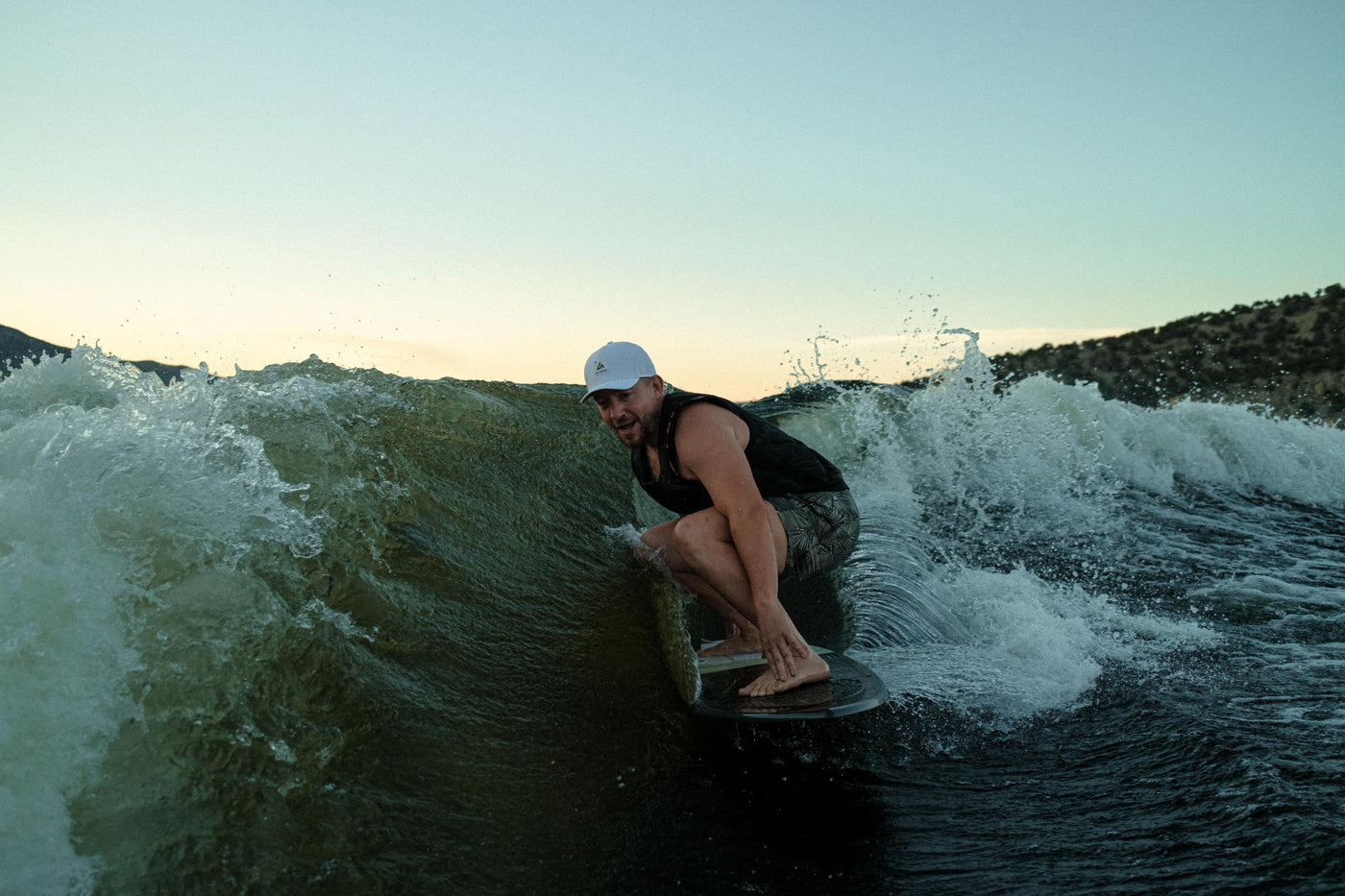 A man in a white Suay Apex hat wakeboards on a dark blue-green lake, skillfully riding a large wave at golden hour with hills in the background.
